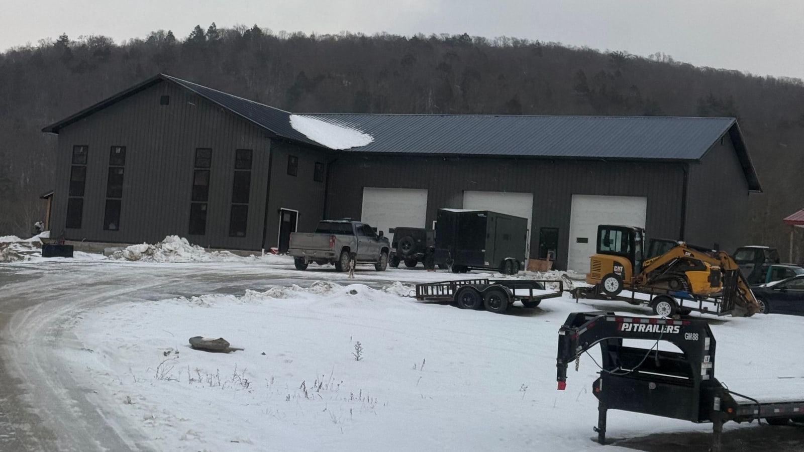 Finished dark metal building with three overhead doors, equipment and trailers parked outside in winter snow